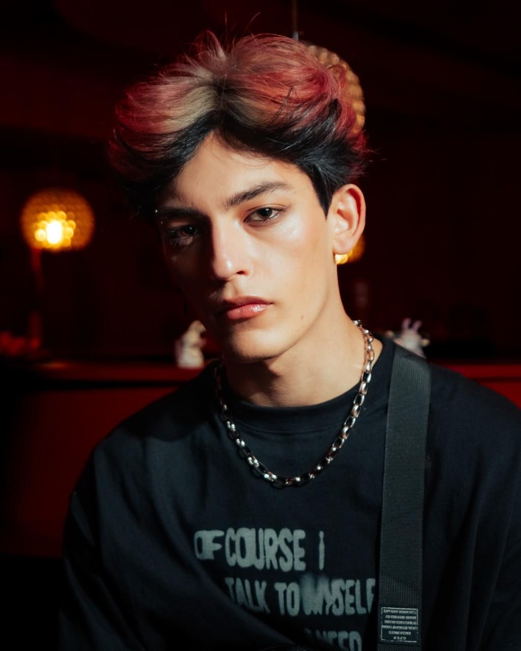 Retrato de una persona de pelo oscuro y un collar de cadena sobre un fondo rojo tenue