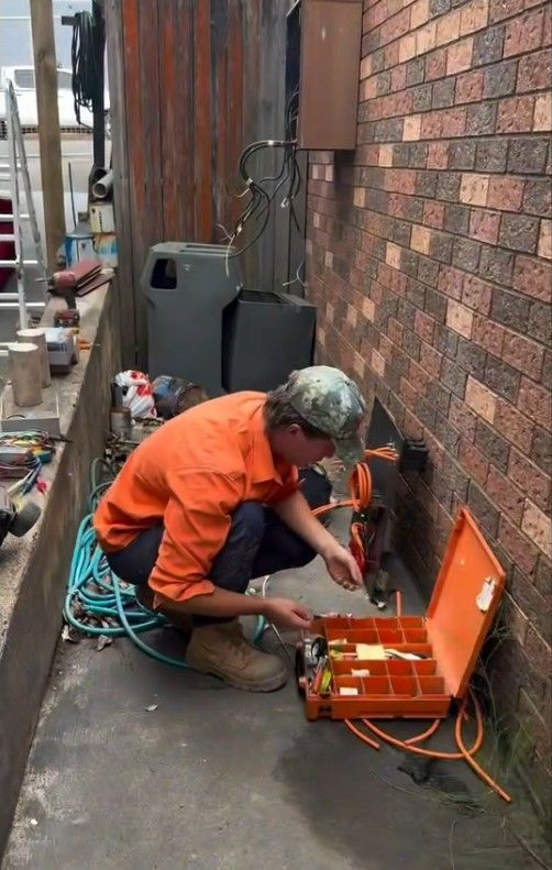 Worker in orange crouches beside brick wall, using tools and an open orange case on a sidewalk. — SRB Electrical Contracting Pty Ltd In Sydney, NSW