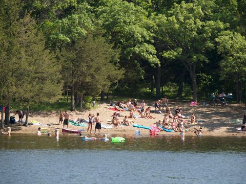 Crowded sandy beach by a lake with swimmers, colorful towels, and green trees in the background