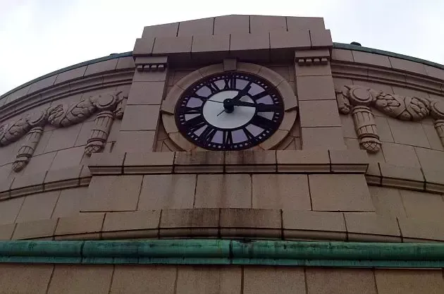 A round analog clock set into a tan, stone-tiled wall with neoclassical decorative carvings and a green metal trim.
