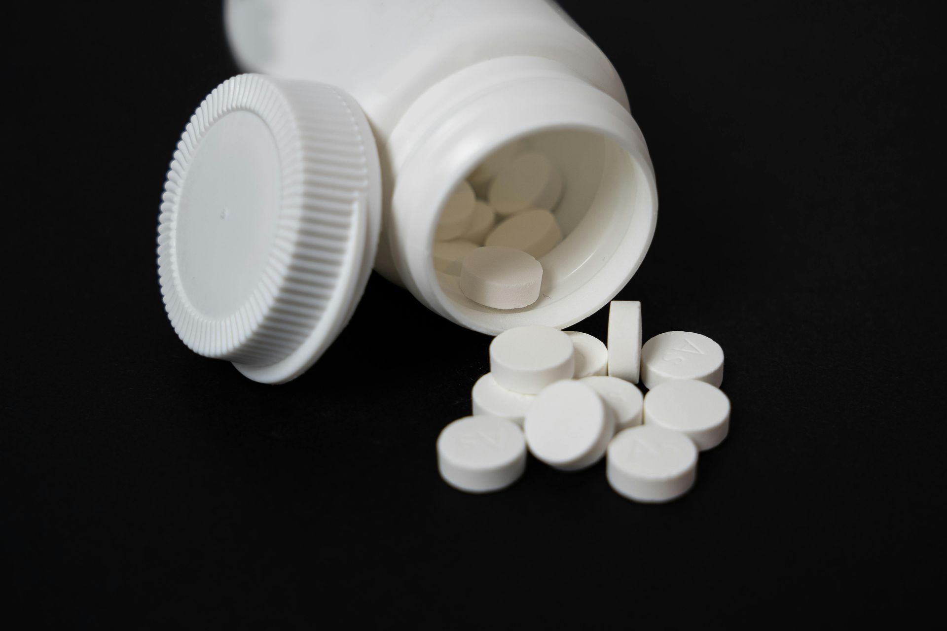 A white plastic pill bottle with its cap lying next to it, surrounded by several scattered white round tablets.