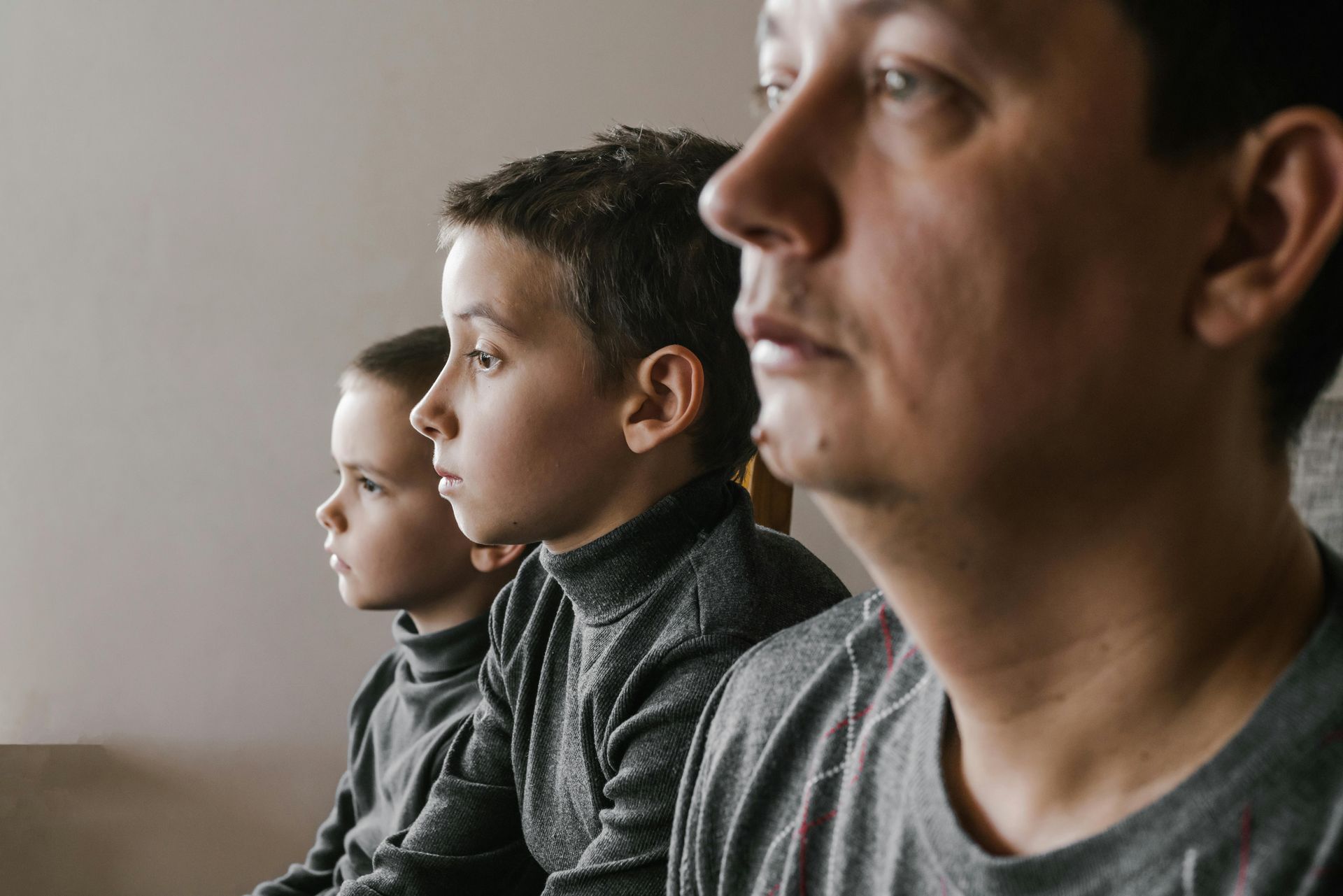 A man and two children sit in a line, looking in the same direction with serious expressions against a plain background.