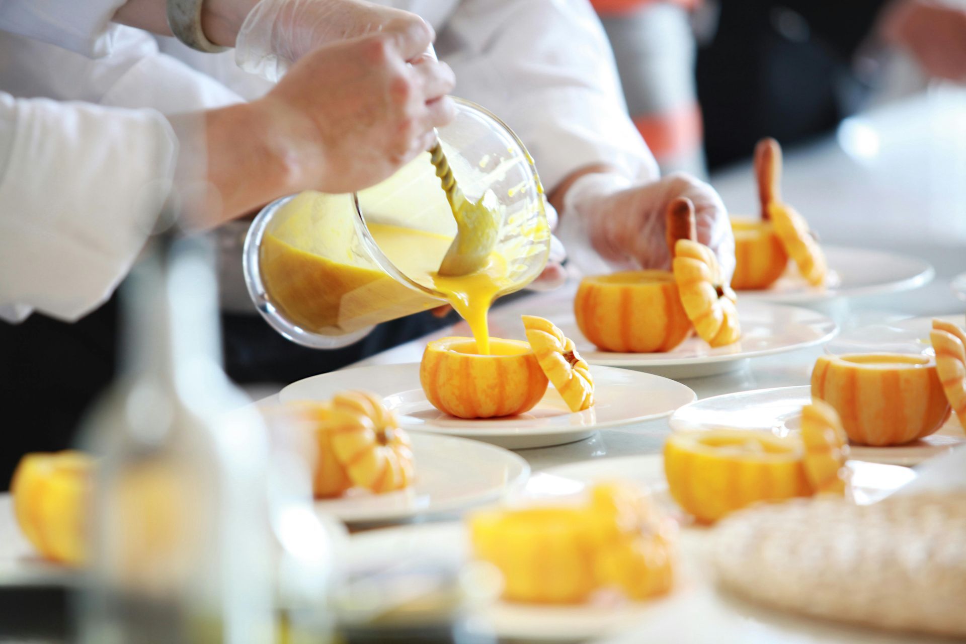 A chef pours golden soup from a glass pitcher into hollowed-out miniature pumpkins arranged on white plates.