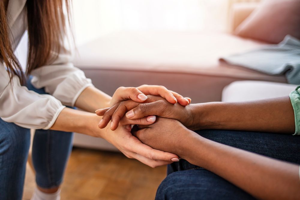 Two people sit holding hands, providing comfort and support in a softly lit, indoor setting.