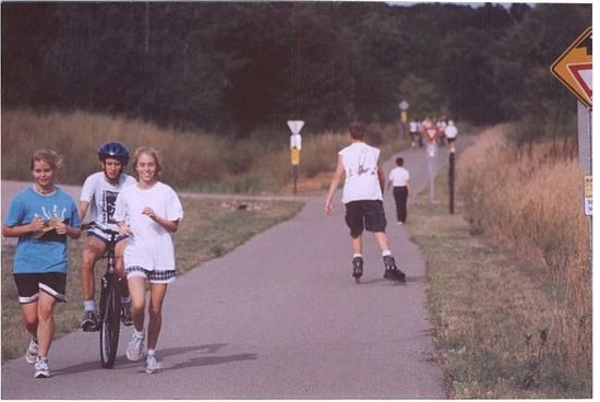 People walking and biking along a paved trail beside tall grass and trees