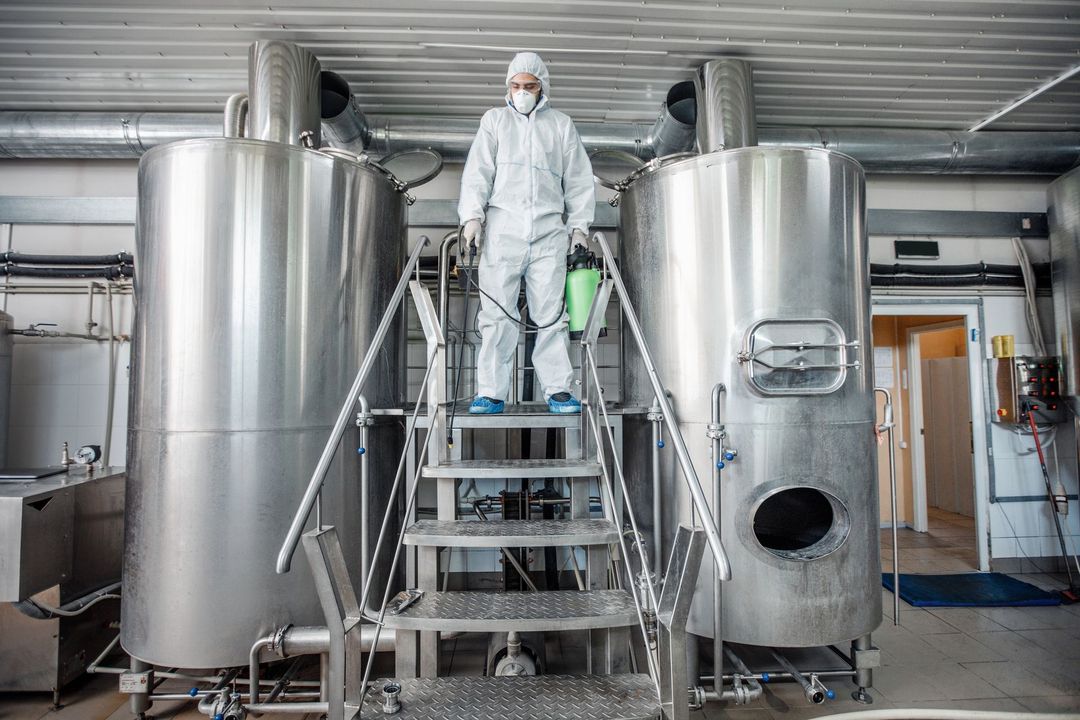Un tecnico con tuta protettiva si trova su una scala metallica tra grandi serbatoi in acciaio inossidabile in un ambiente industriale pulito.