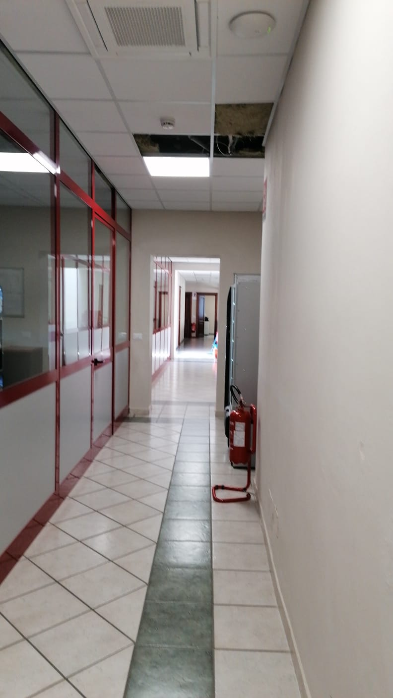 Stretto corridoio d'ufficio con pavimento piastrellato, pareti in vetro con cornice rossa e un estintore sulla destra.