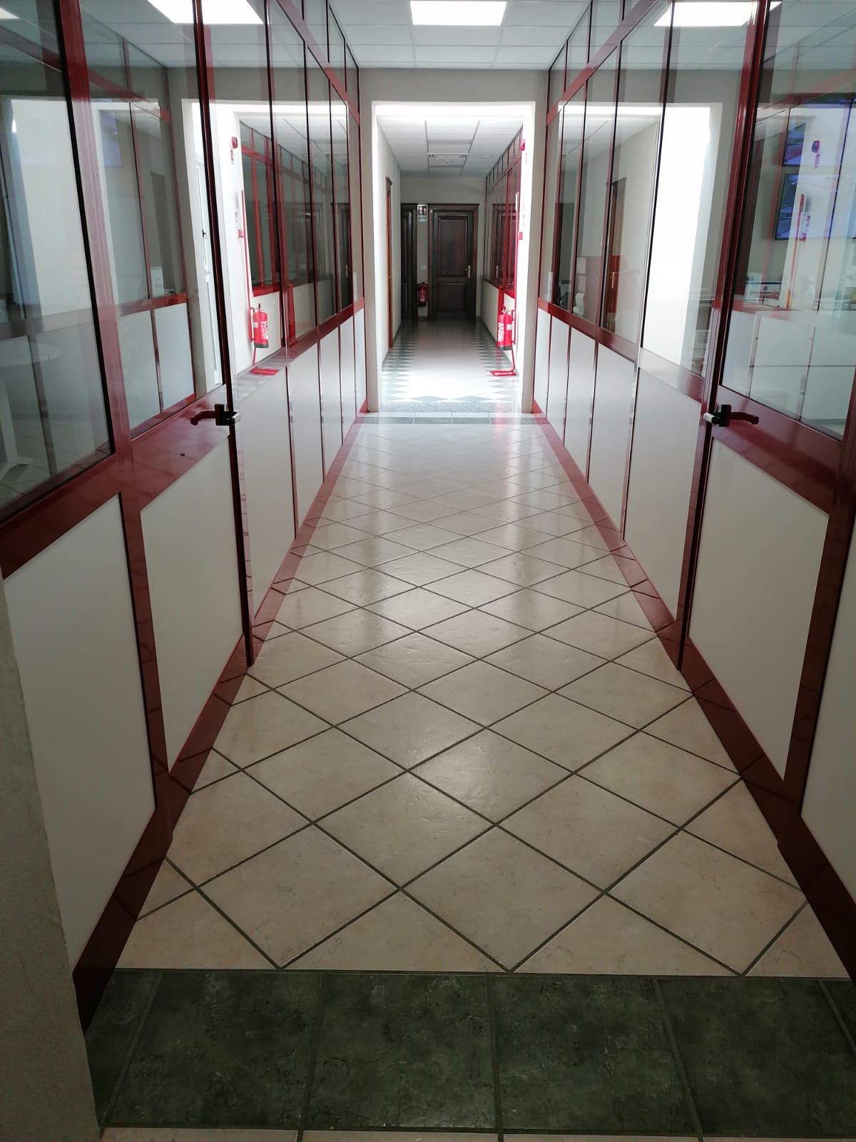 Corridoio vuoto con pareti di vetro incorniciate di rosso e pavimento piastrellato, che conduce a una porta luminosa
