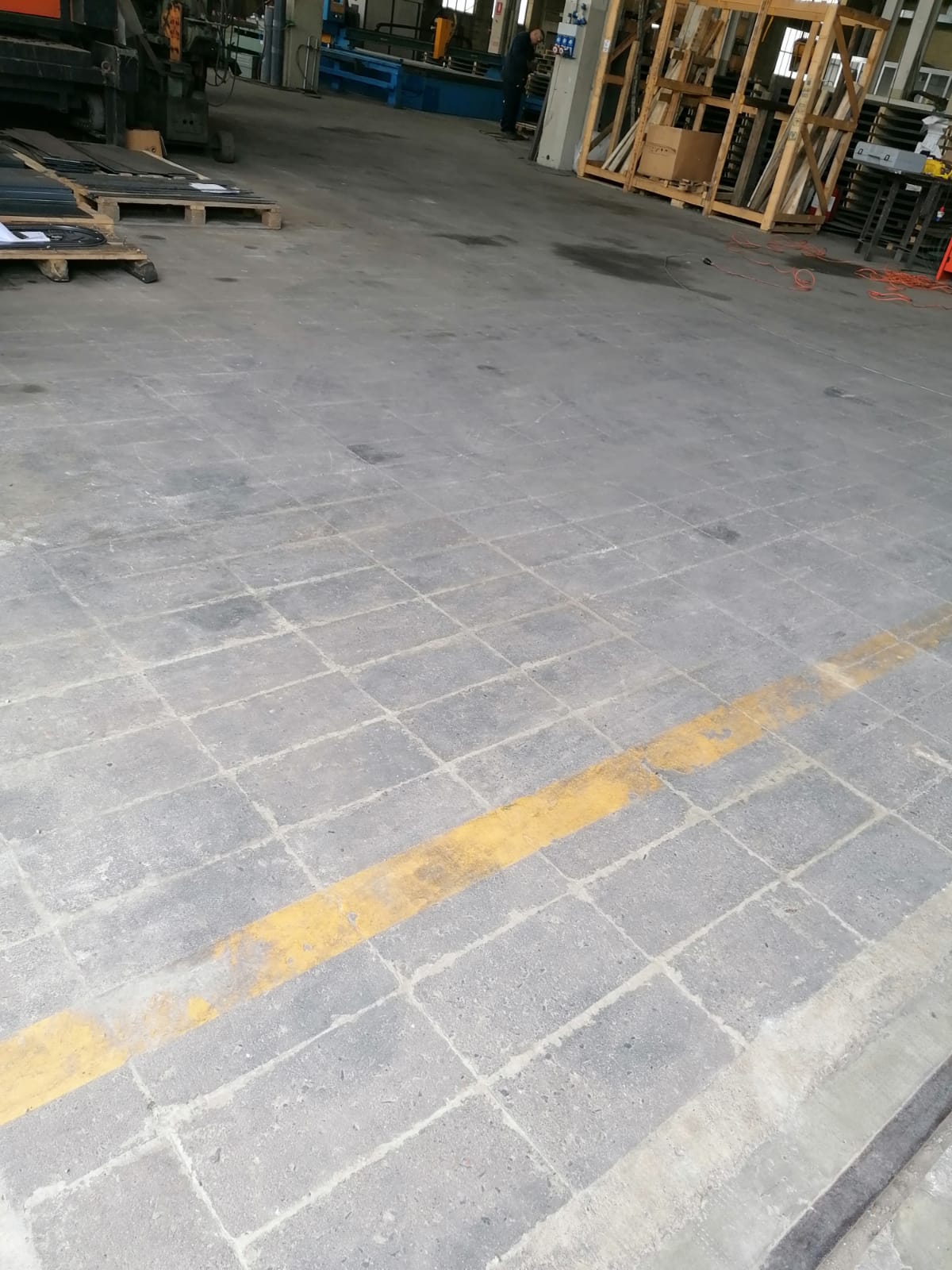 Pavimento di un magazzino con una linea gialla, pallet e materiali impilati sullo sfondo.