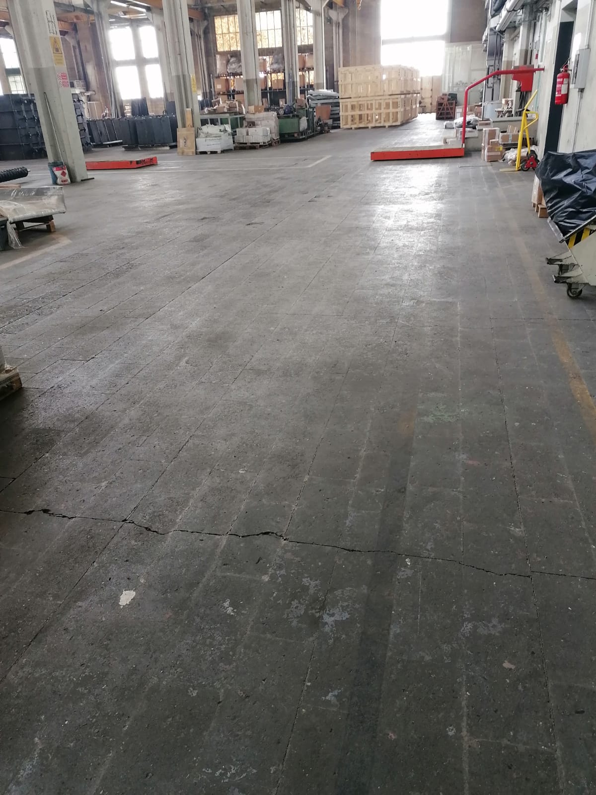 Magazzino industriale vuoto con pavimento in cemento, finestre alte e attrezzature sparse ai margini.
