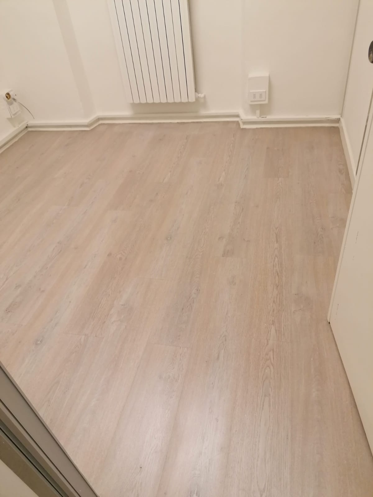 Piccola stanza vuota con pavimento in legno chiaro, pareti bianche e un termosifone sotto la finestra.
