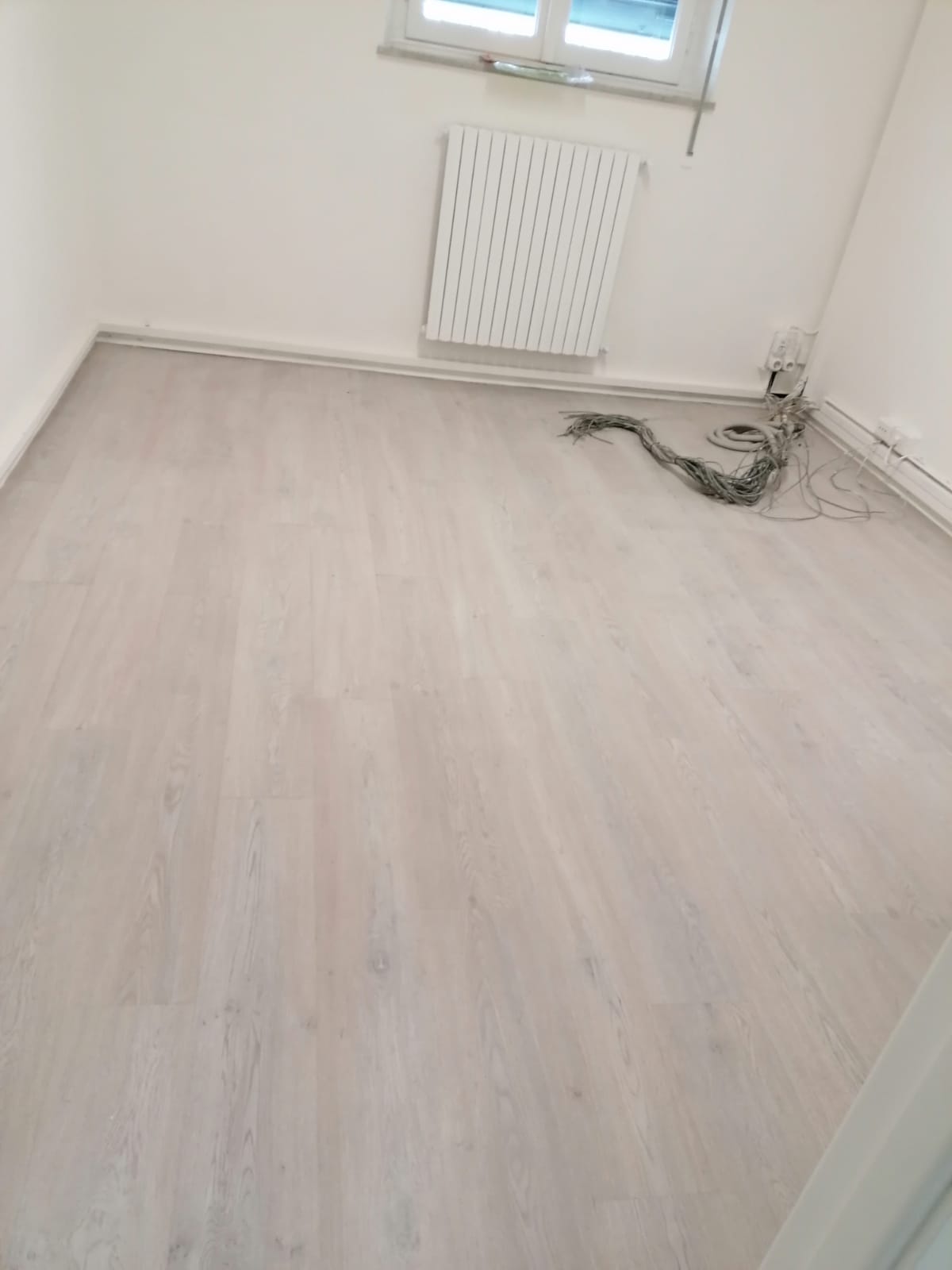 Stanza vuota con pavimento in legno chiaro, pareti bianche, finestra, termosifone e cavi in ​​un angolo.