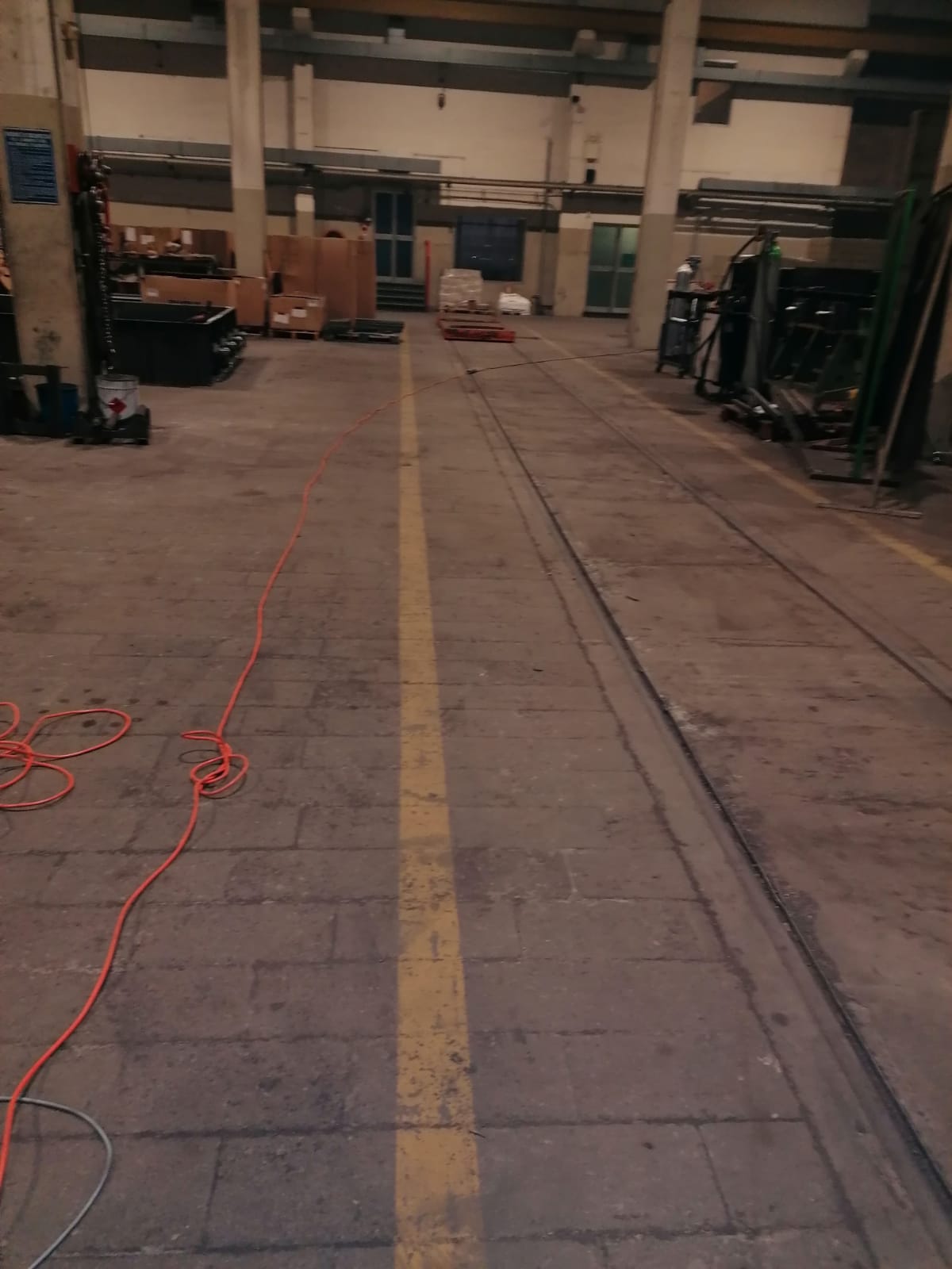 Corridoio vuoto di un magazzino con una linea gialla al centro e cavi sparsi sul pavimento di cemento.