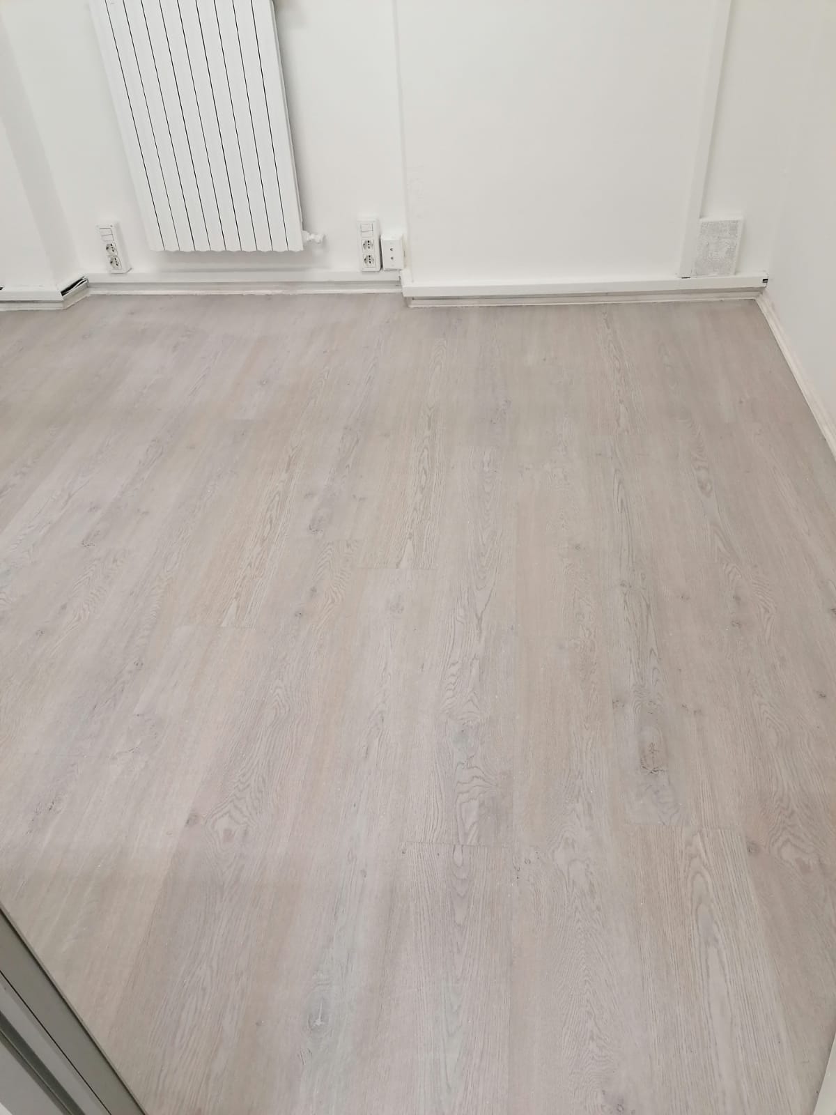 Stanza vuota con pavimento in legno chiaro e pareti bianche, mostrata vicino a un termosifone.