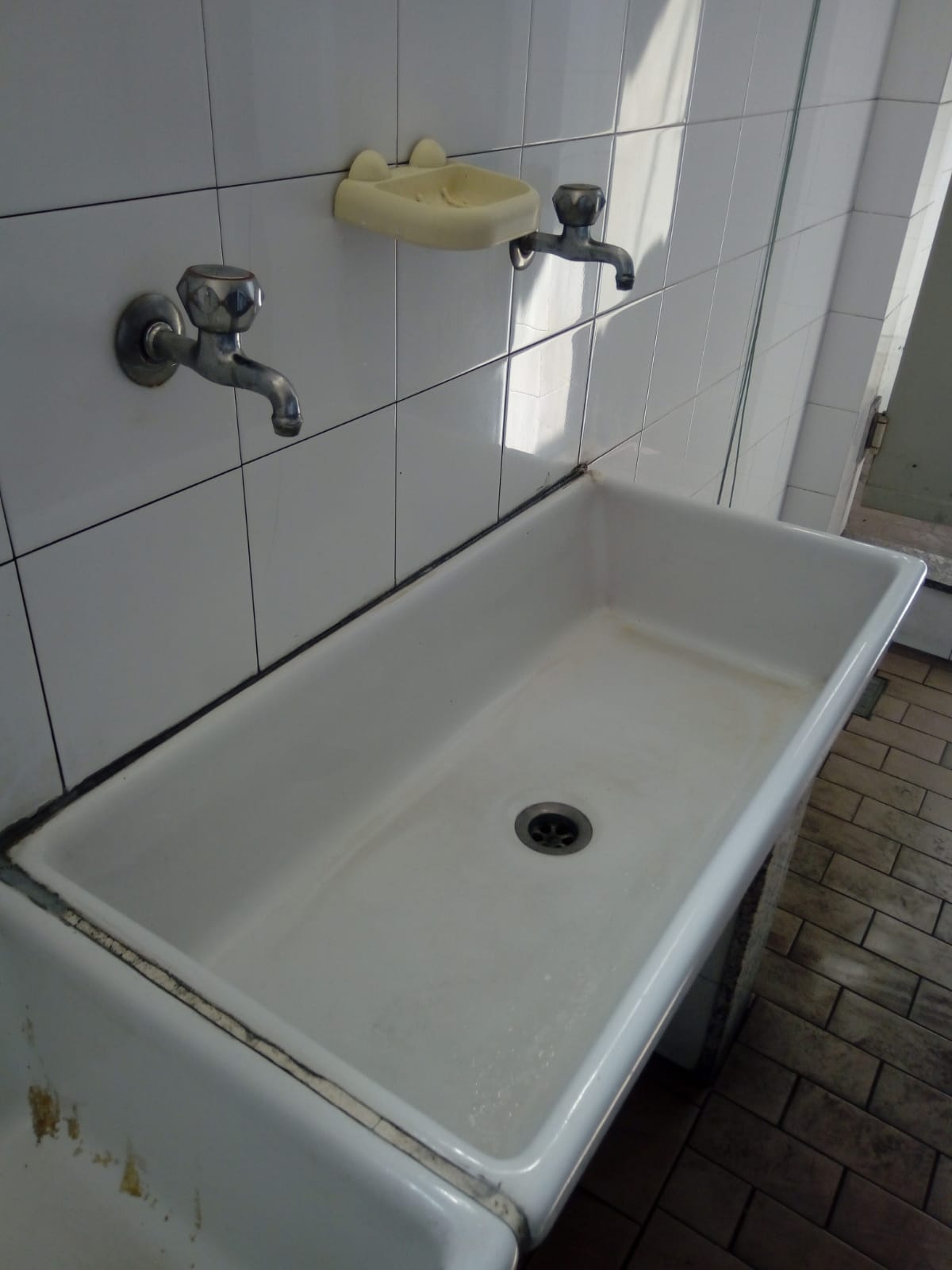 Vecchio lavabo di servizio bianco con due rubinetti in un bagno o una toilette piastrellata.