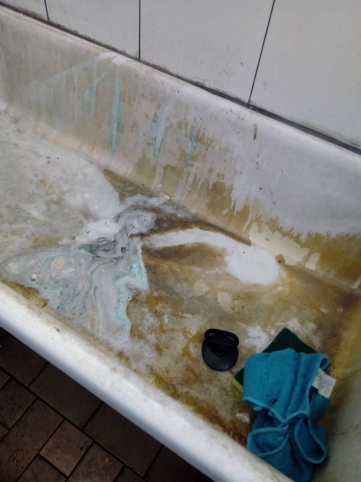 Vasca da bagno macchiata con acqua fangosa, tappo nero e un panno blu piegato sul bordo.