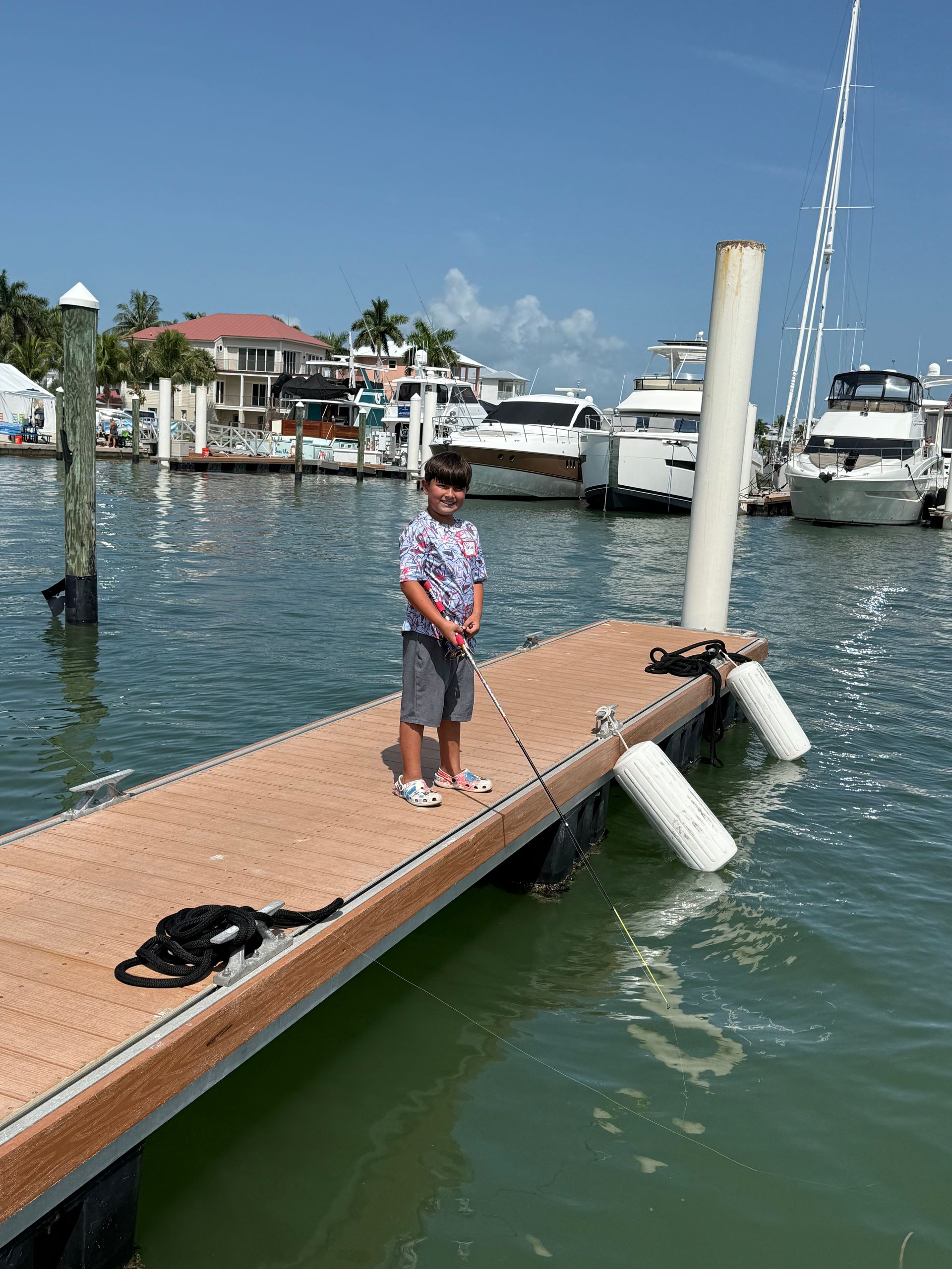 Child standing on a dock by boats in a marina, holding a pole on a sunny day