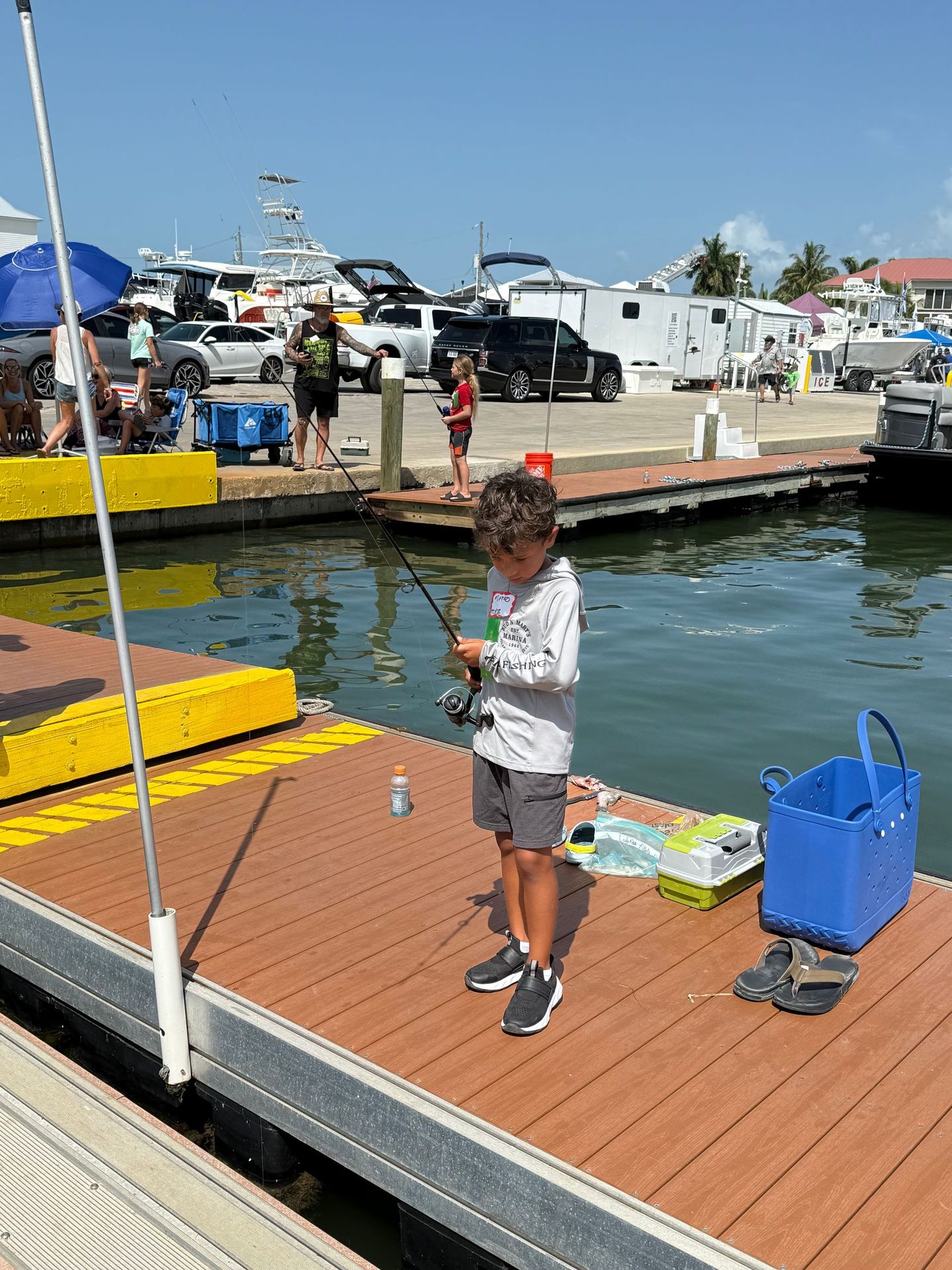 Child fishing from a dock beside a marina, with blue bucket and bright yellow boats in the background