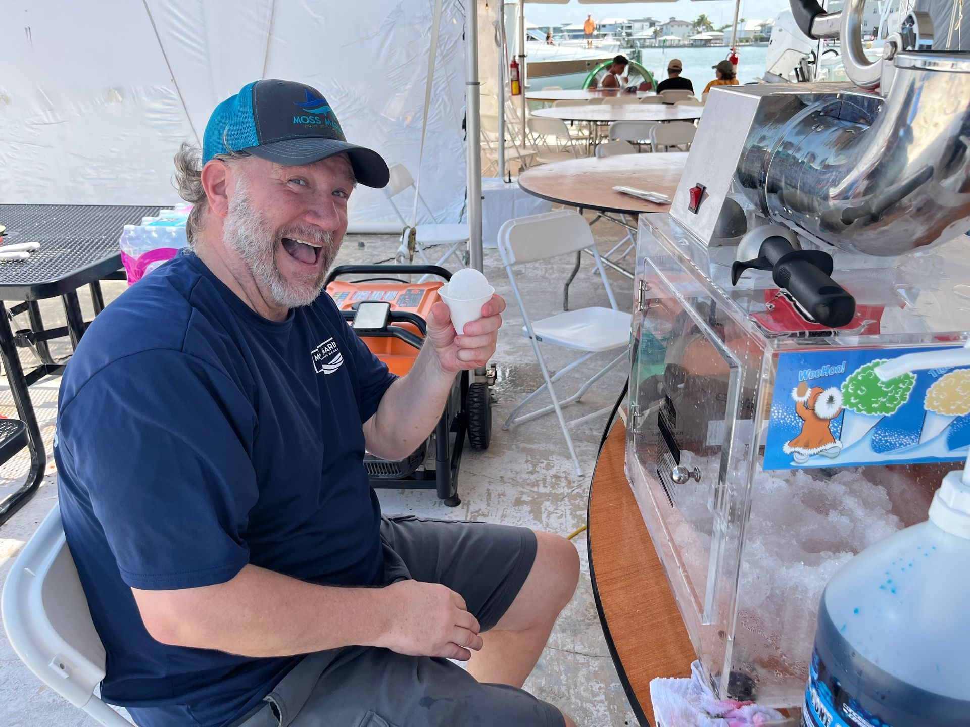 Smiling man in a navy shirt holding a drink at a marina table with ice and boats in the background