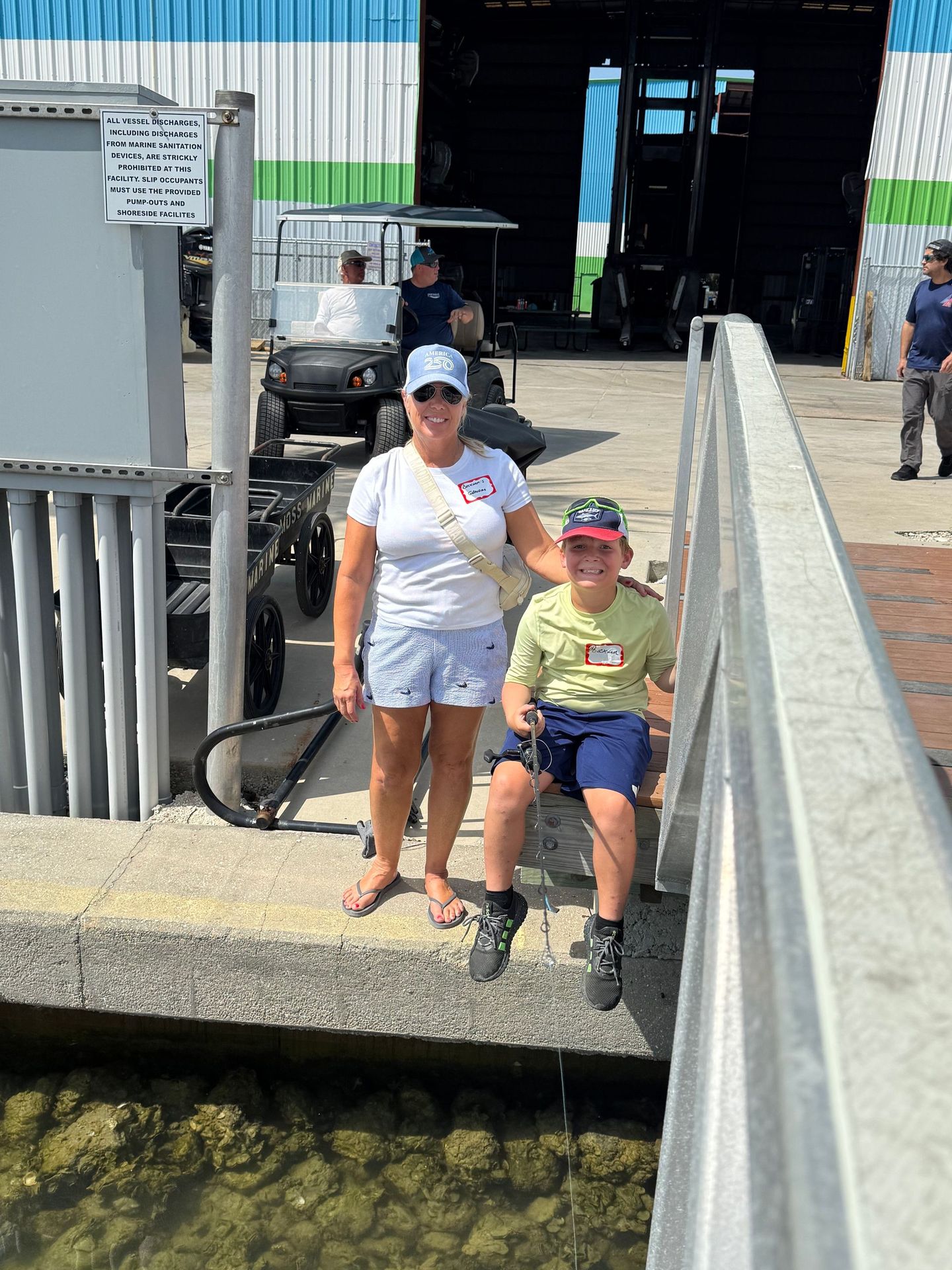 Woman and child posing by a waterfront dock in sunlight, with boats and a warehouse behind them