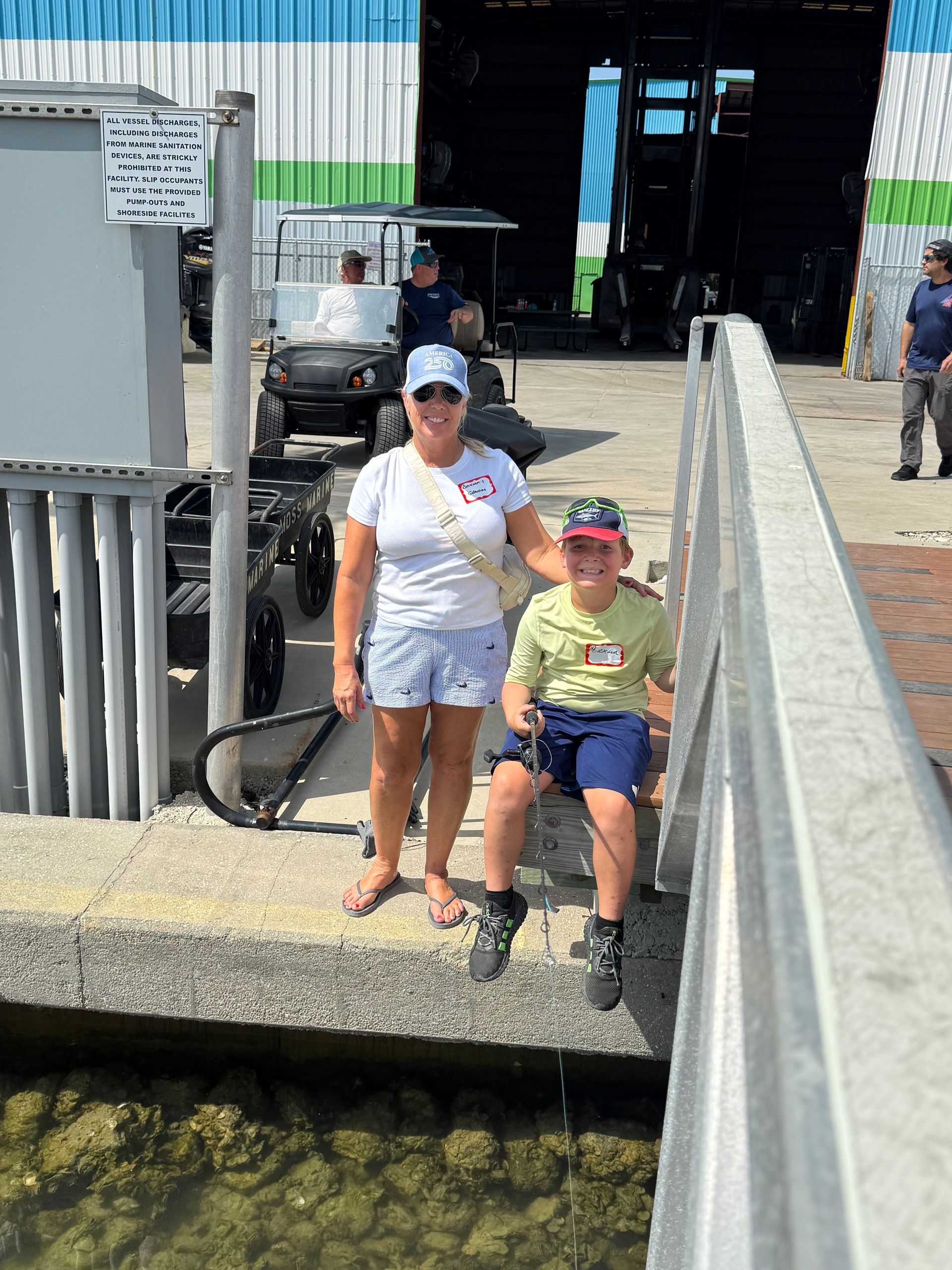 Woman and child posing by a waterfront dock in sunlight, with boats and a warehouse behind them