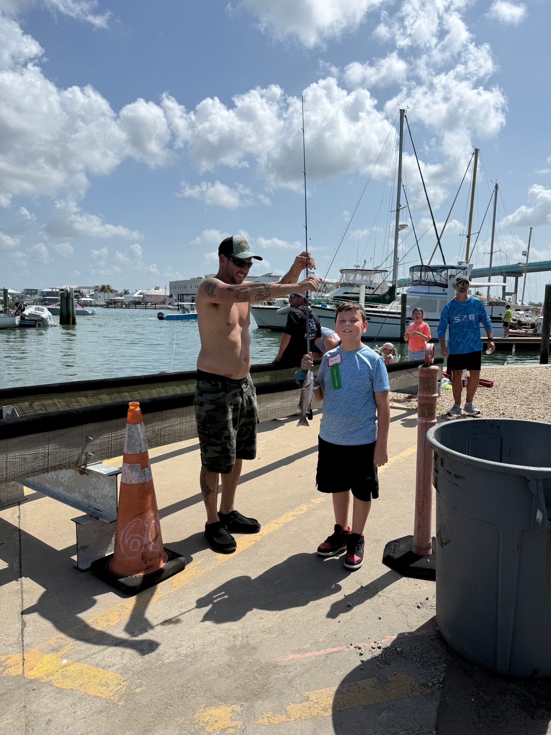 Two people posing by a harbor with boats, orange cones, and a blue sky with clouds.