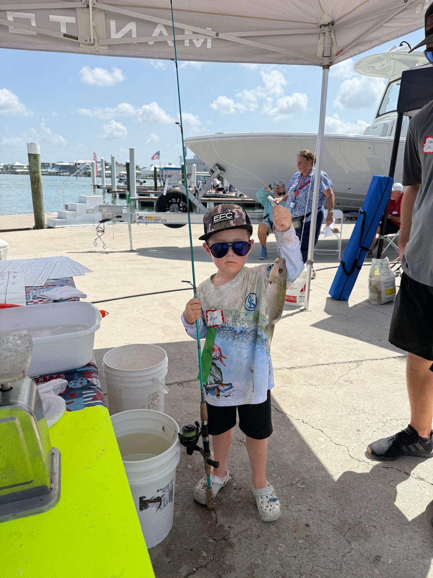 Boy holding a fish at a sunny dockside event under a canopy, with buckets and people nearby.