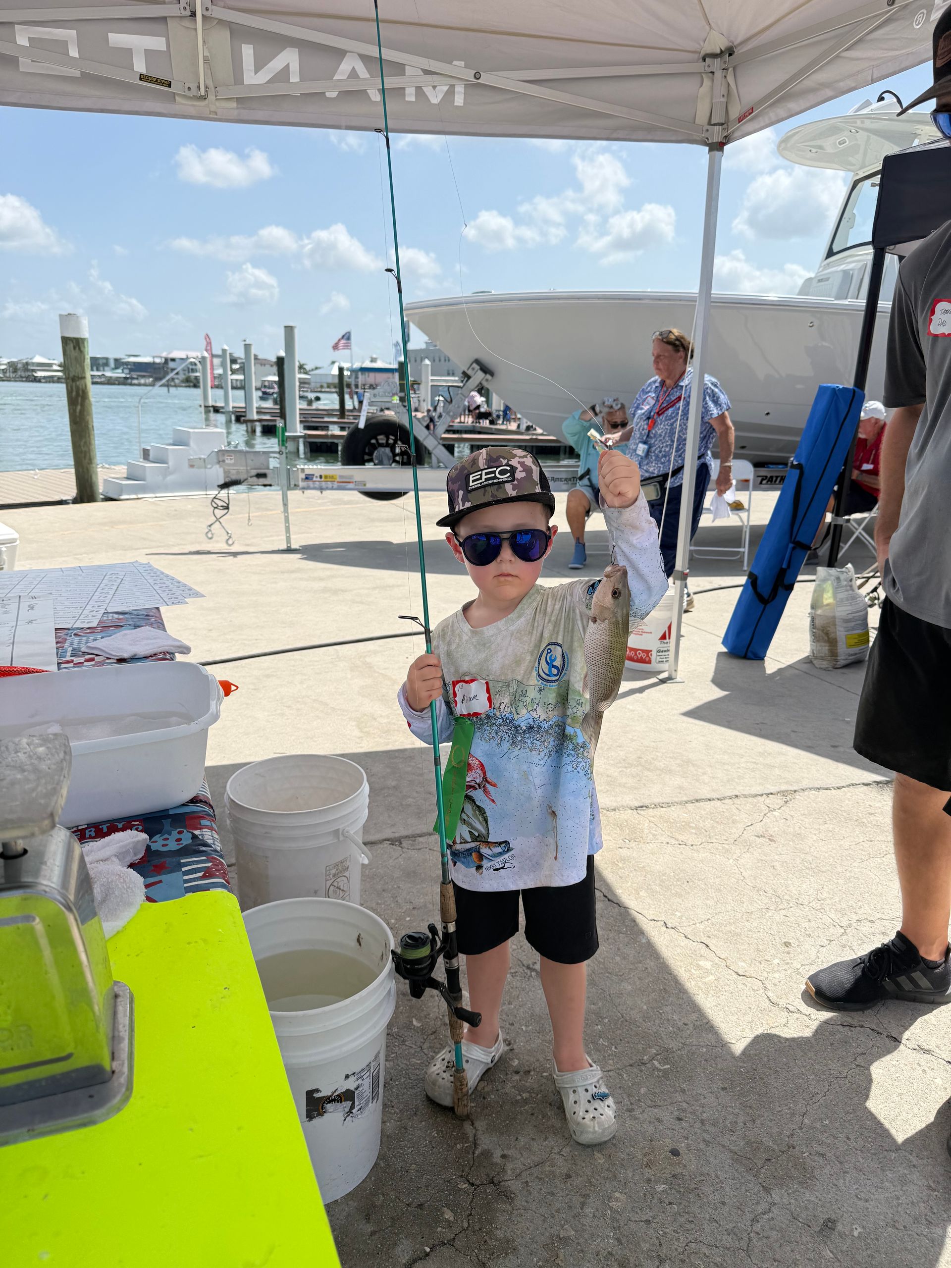 Boy holding a fish at a sunny dockside event under a canopy, with buckets and people nearby.