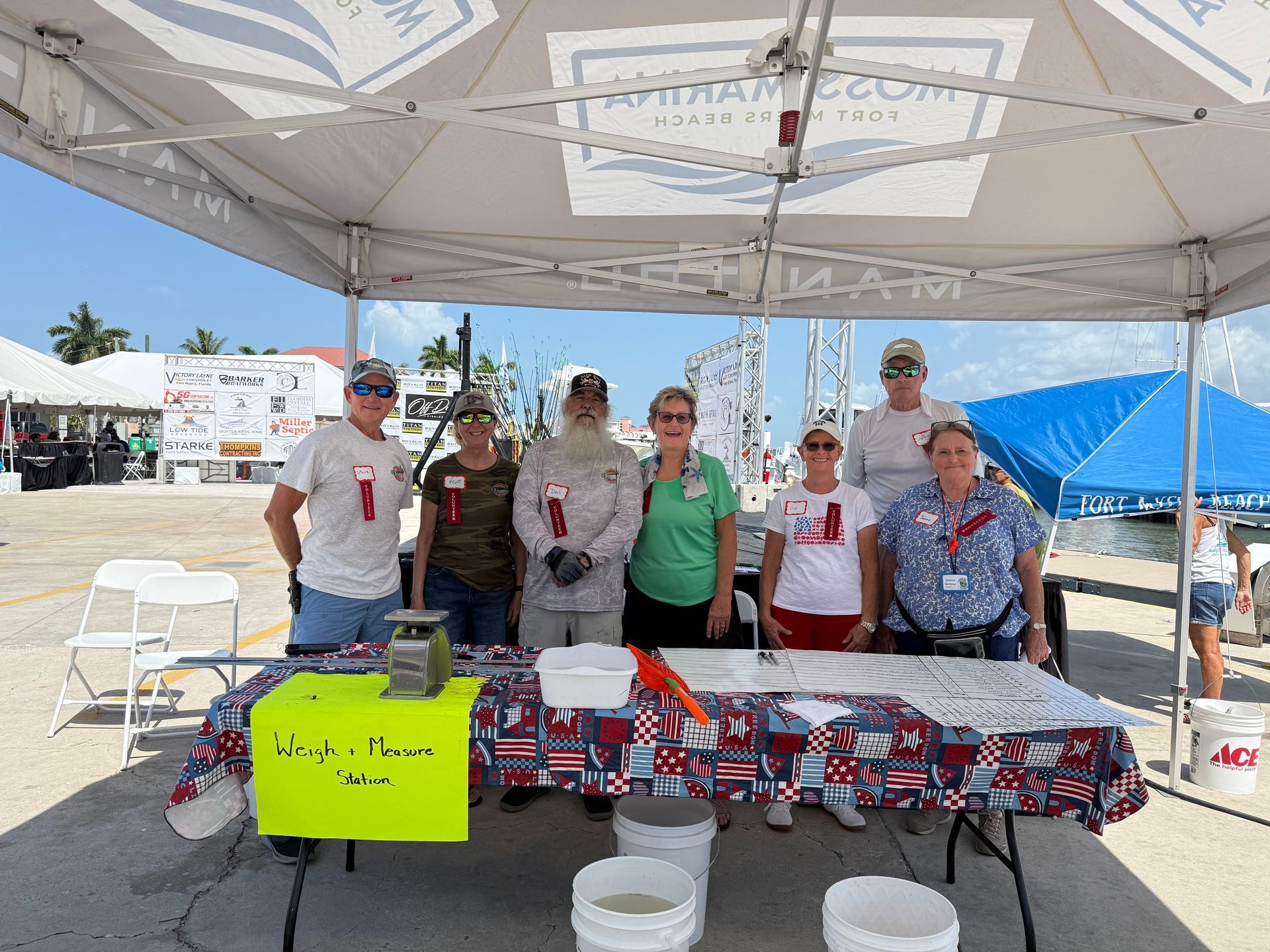 Six volunteers stand behind a patriotic table display under a white canopy at an outdoor event.