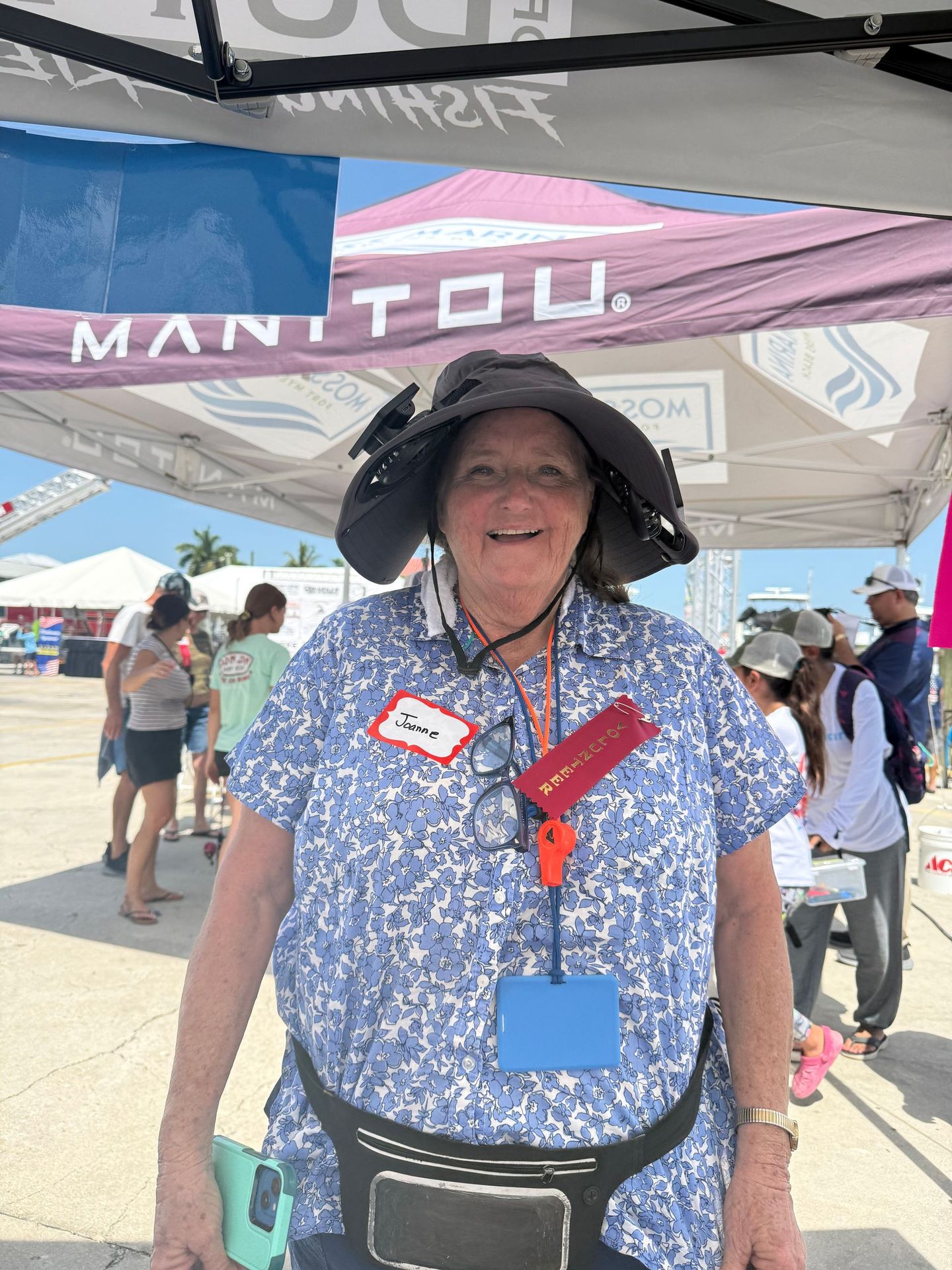 Smiling person in blue floral shirt and sun hat under a MANTOU tent at an outdoor event