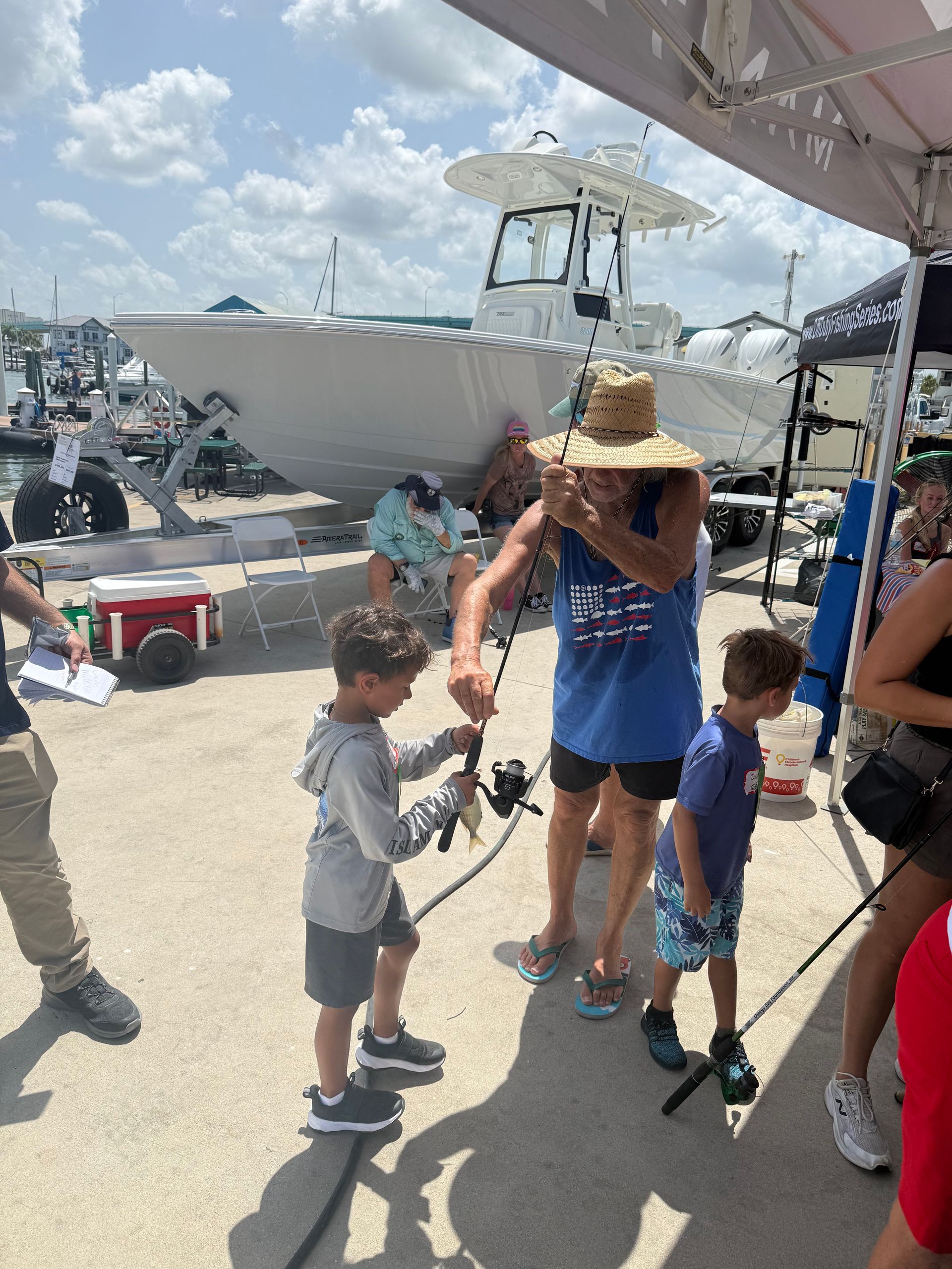 Person in blue shirt and straw hat stands on a dock by a boat while a child reaches up holding a camera tripod