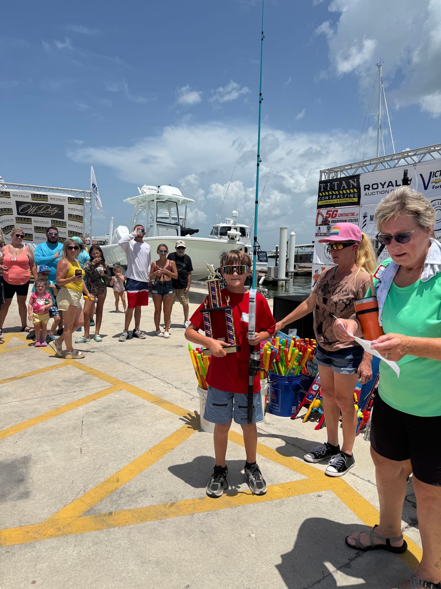 People gathered on a sunny dockside, with children and adults standing around a red-shirted boy holding a microphone.