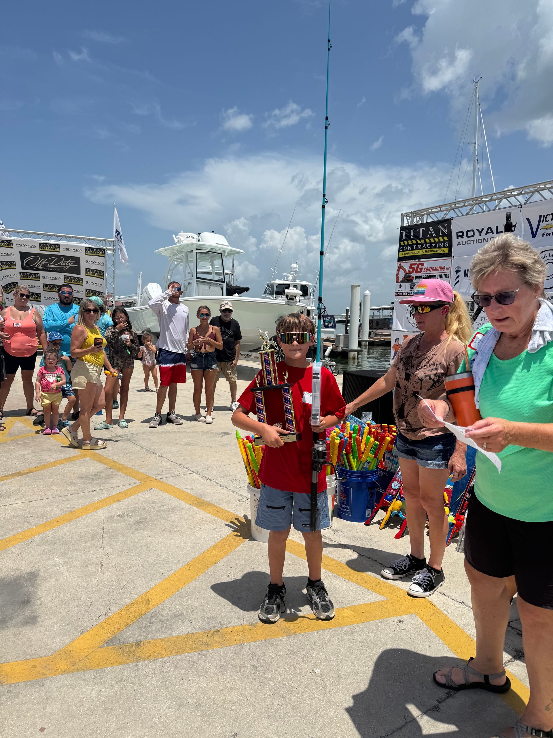 People gathered on a sunny dockside, with children and adults standing around a red-shirted boy holding a microphone.