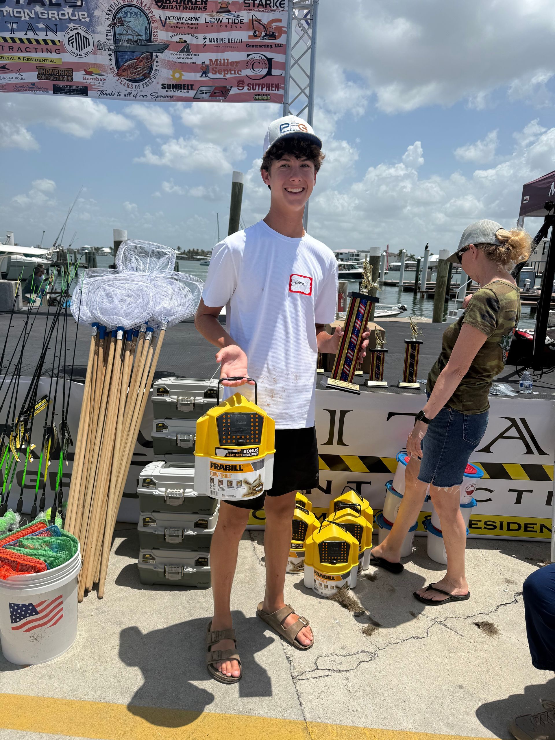 Person holding yellow device at a waterfront event, with other people and signs in the background.