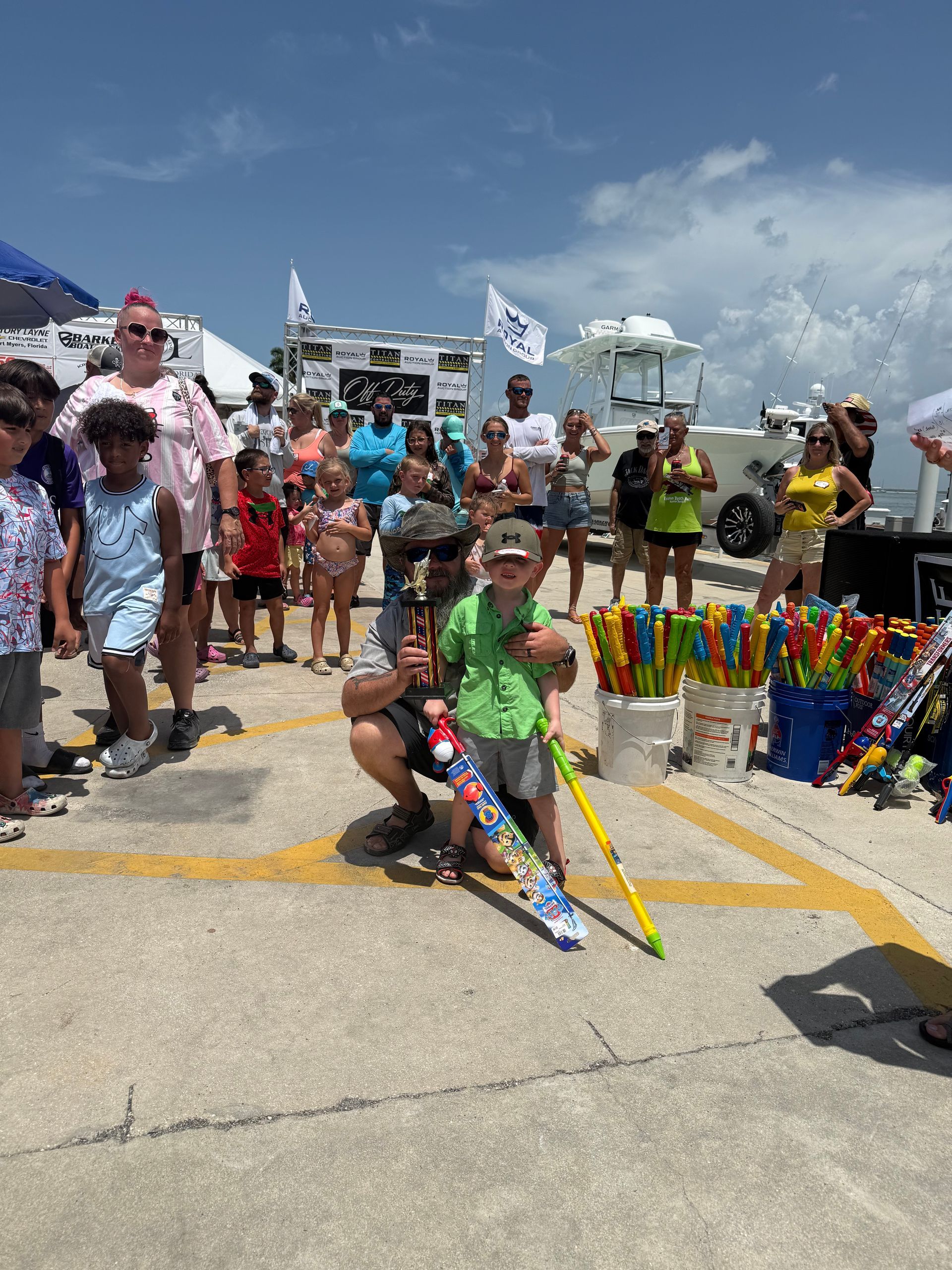 Crowd at a sunny beach event with a man kneeling beside colorful umbrellas and a kite display