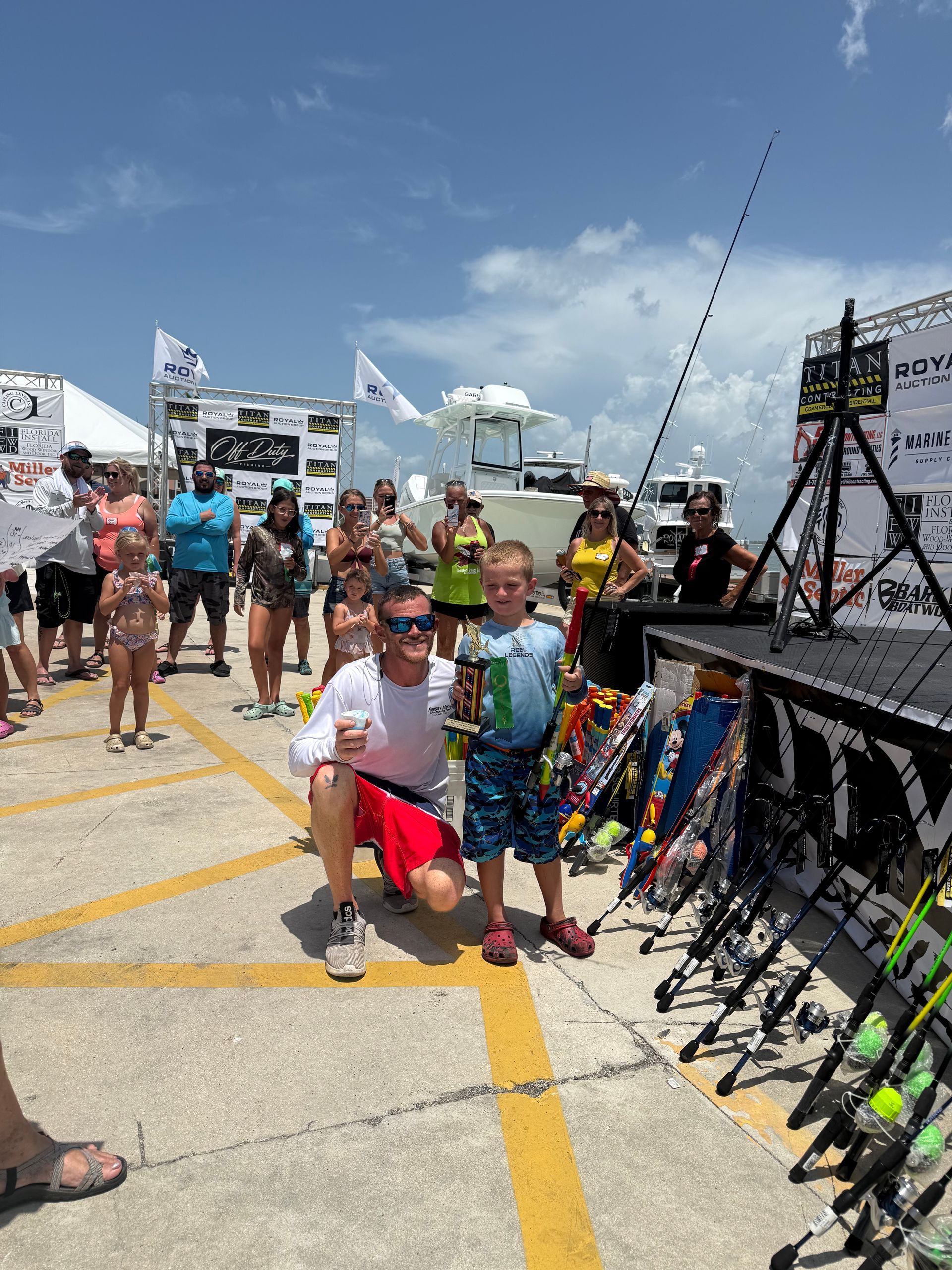 Two people posing on a dock at a fishing tournament, with fishing rods and spectators in the background.