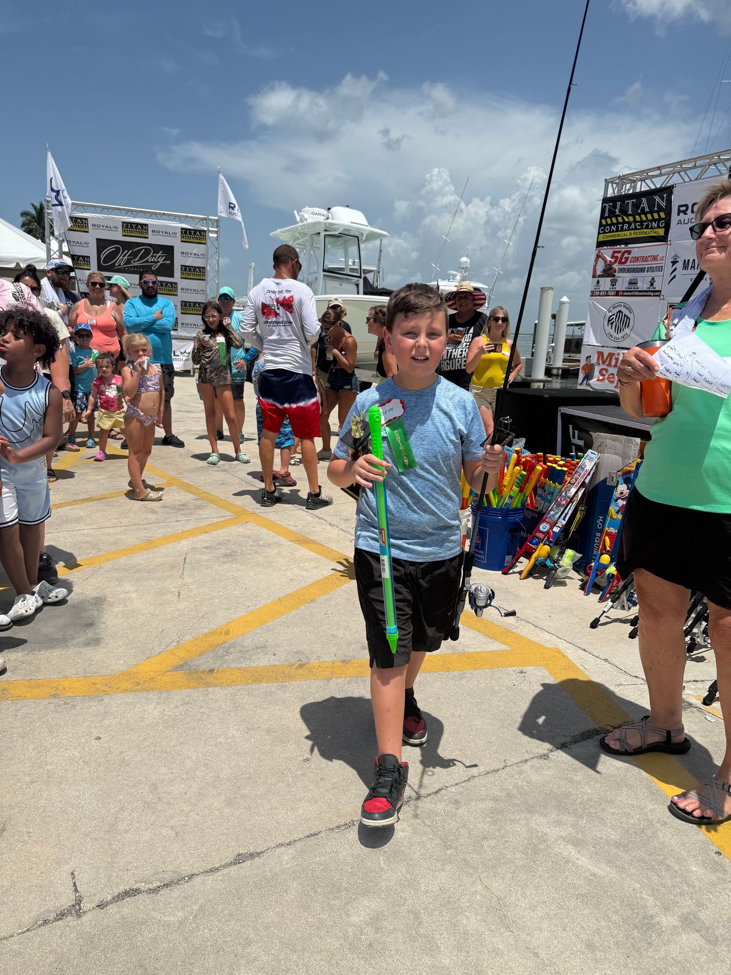 Boy holding a green balloon toy at an outdoor event, with other people and booths in the background