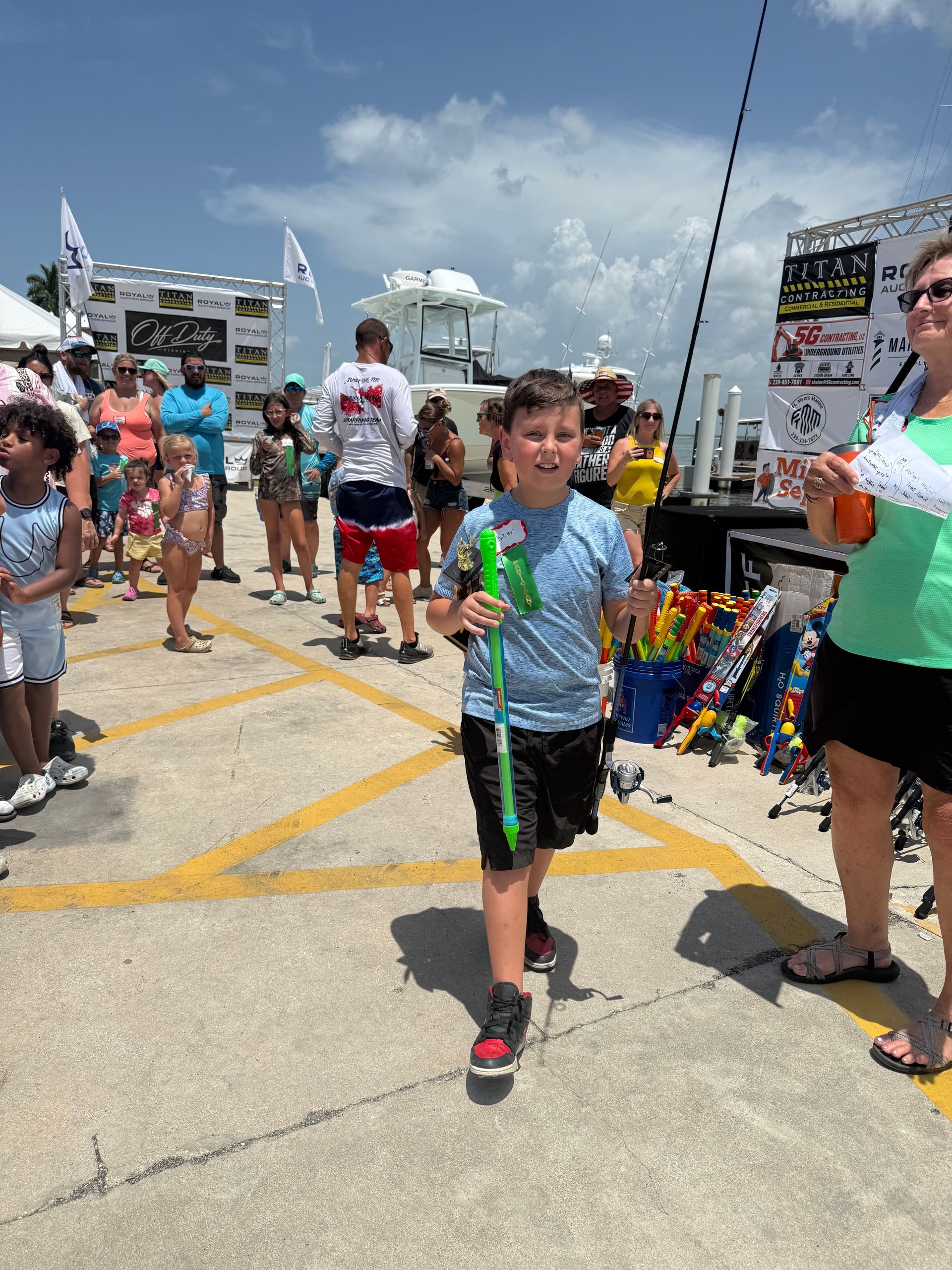 Boy holding a green balloon toy at an outdoor event, with other people and booths in the background