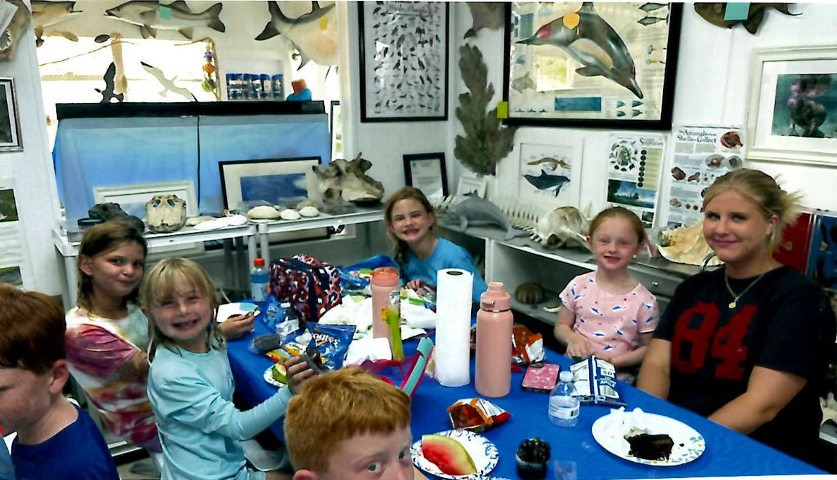 Children sitting around a blue table at a classroom snack or party, smiling with drinks and snacks.