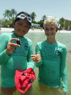 Two swimmers in green rash guards holding seashells at a sunny beach, smiling at the camera
