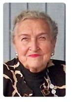 An older woman with short gray hair smiles, wearing a black and brown patterned top and jewelry.