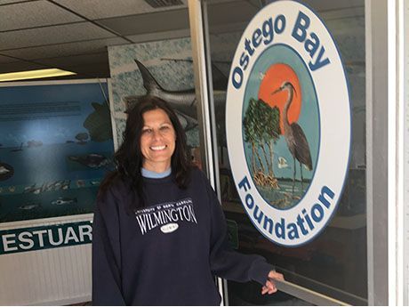 Woman standing by Oswego Bay Foundation sign at an estuary office, smiling and pointing.