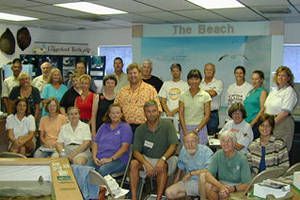 Group of people posing indoors in a room labeled “The Beach,” with a blue mural and office supplies in front.