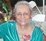 Elderly woman with gray hair wearing glasses and a teal top, smiling, outdoors.