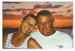 Smiling couple embracing at sunset. Woman in striped top, man in white shirt. Orange sky and water in background.