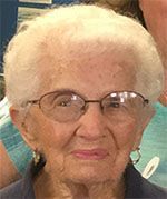 An elderly woman with white hair and glasses smiles slightly. She wears hoop earrings and a navy top.