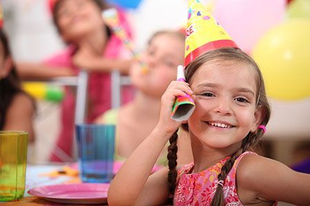 Girl in a party hat smiles, holding a party horn at a birthday party. Other children and balloons are in the background.