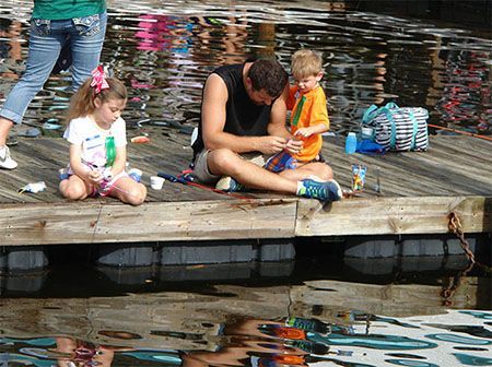 Children sitting on a wooden dock by the water, one man helping a child with something in hand.