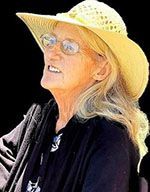 Woman with blonde hair wearing a yellow sun hat and glasses, looking upwards. She is wearing a black shirt.