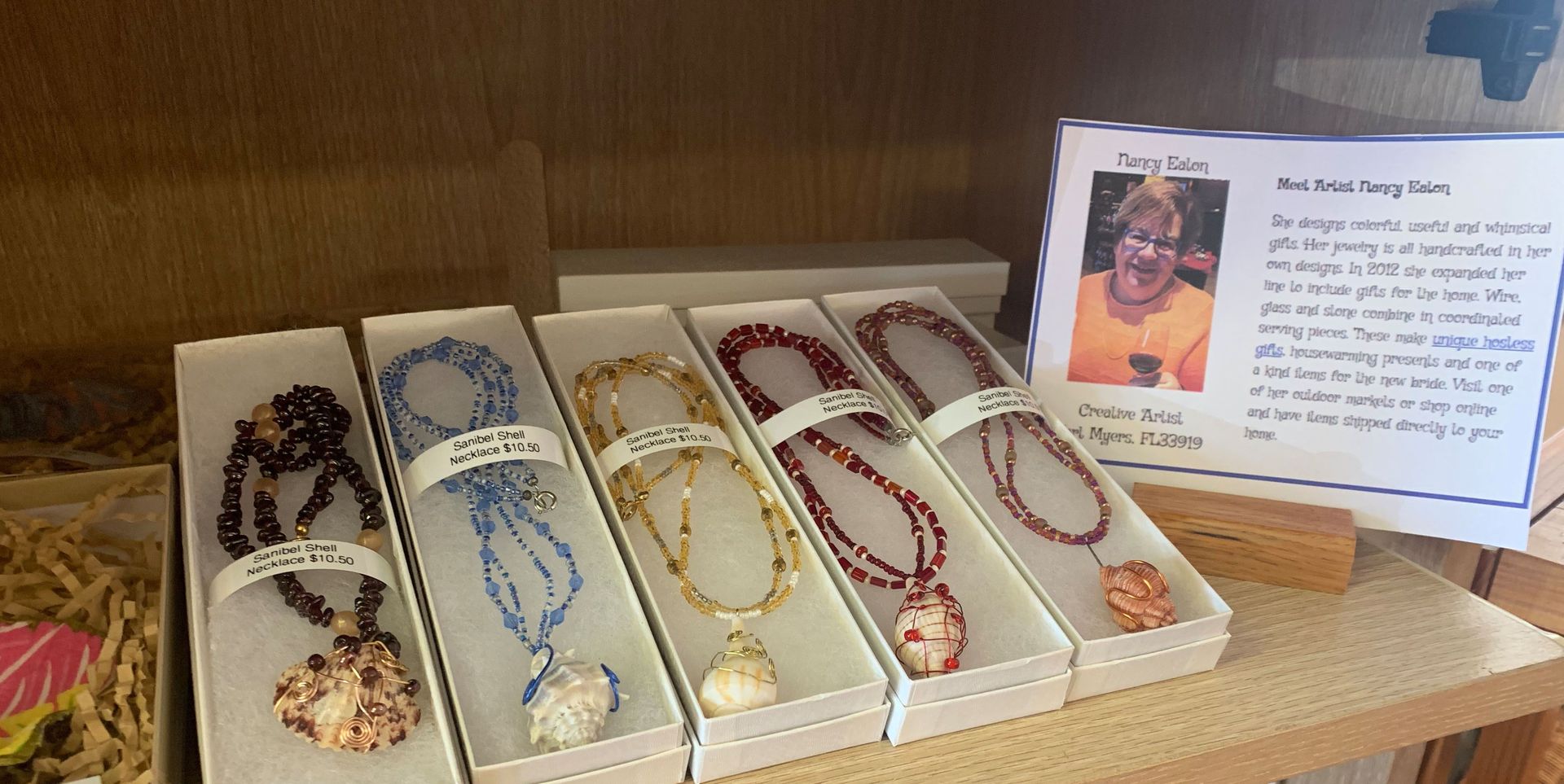 Necklaces with seashell pendants in white boxes on display, next to a sign with a photo and text.